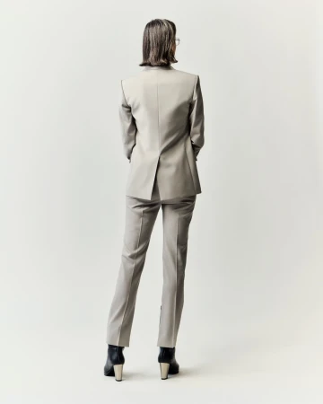 Rear view of an adult woman in a light grey suit with black boots, standing against a white backdrop, showcasing formal business attire.