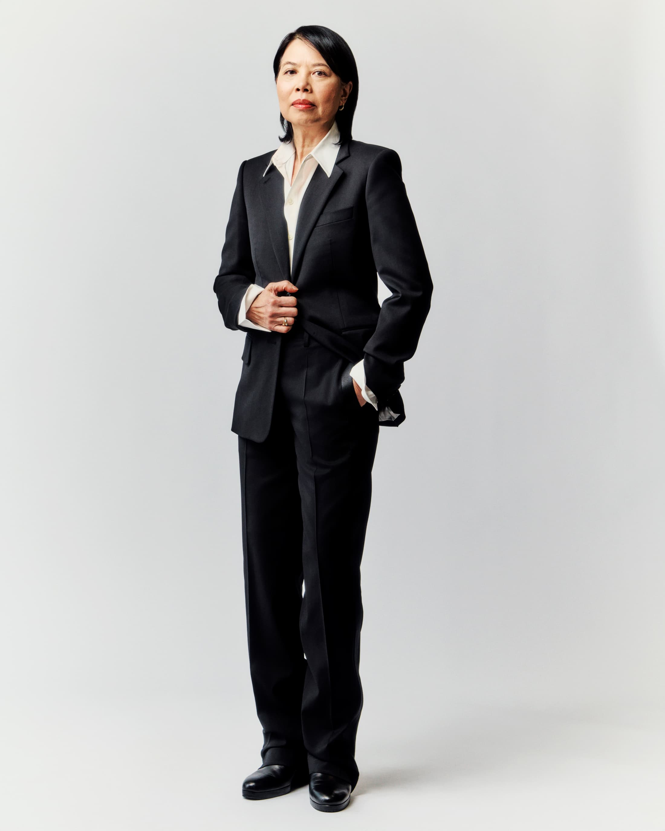 Full-length portrait of an Asian woman in a black pantsuit and white shirt, standing against a plain, light gray background in a photo studio.