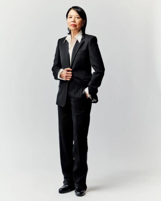 Full-length portrait of an Asian woman in a black pantsuit and white shirt, standing against a plain, light gray background in a photo studio.
