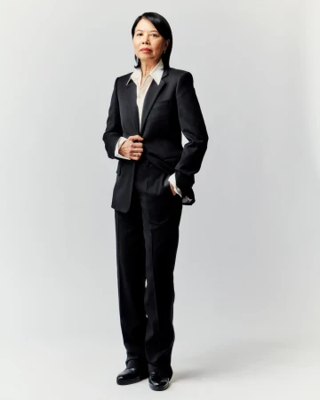 Full-length portrait of an Asian woman in a black pantsuit and white shirt, standing against a plain, light gray background in a photo studio.