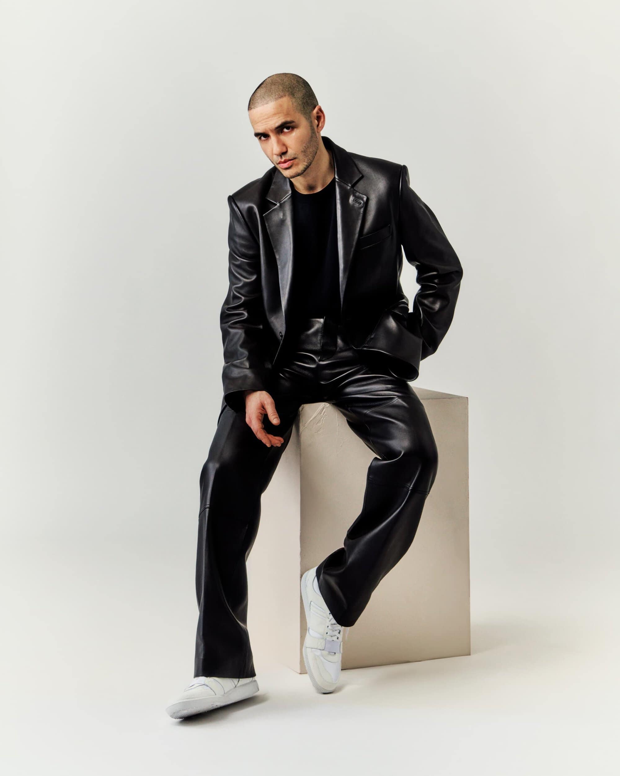 An adult male is sitting on a white cube wearing a shiny black blazer, trousers, and white sneakers in a photo studio.