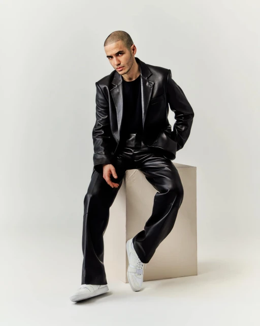 An adult male is sitting on a white cube wearing a shiny black blazer, trousers, and white sneakers in a photo studio.