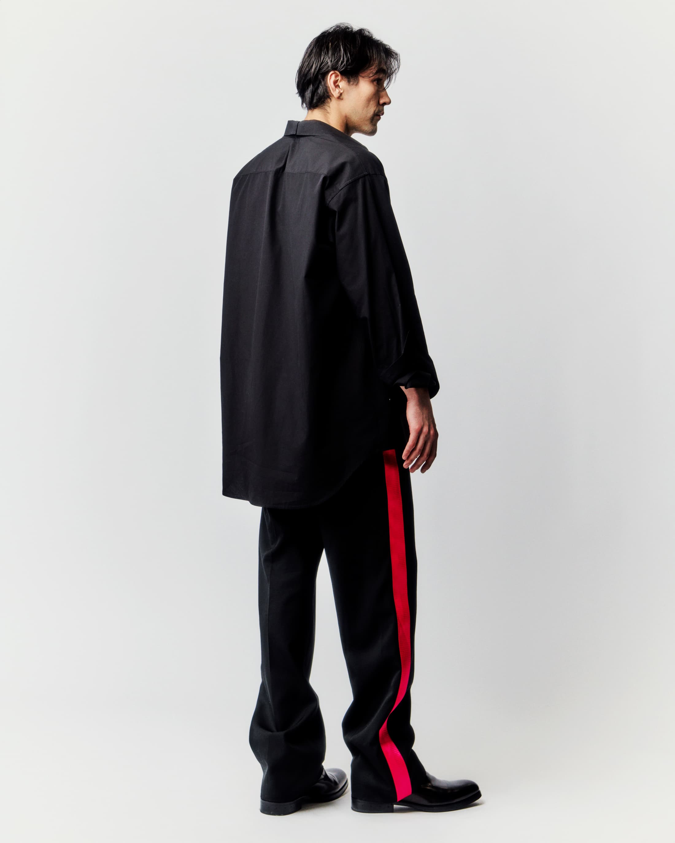 Back view of an adult male model in an oversized black button-down shirt and black pants with a red stripe, standing in a photo studio.