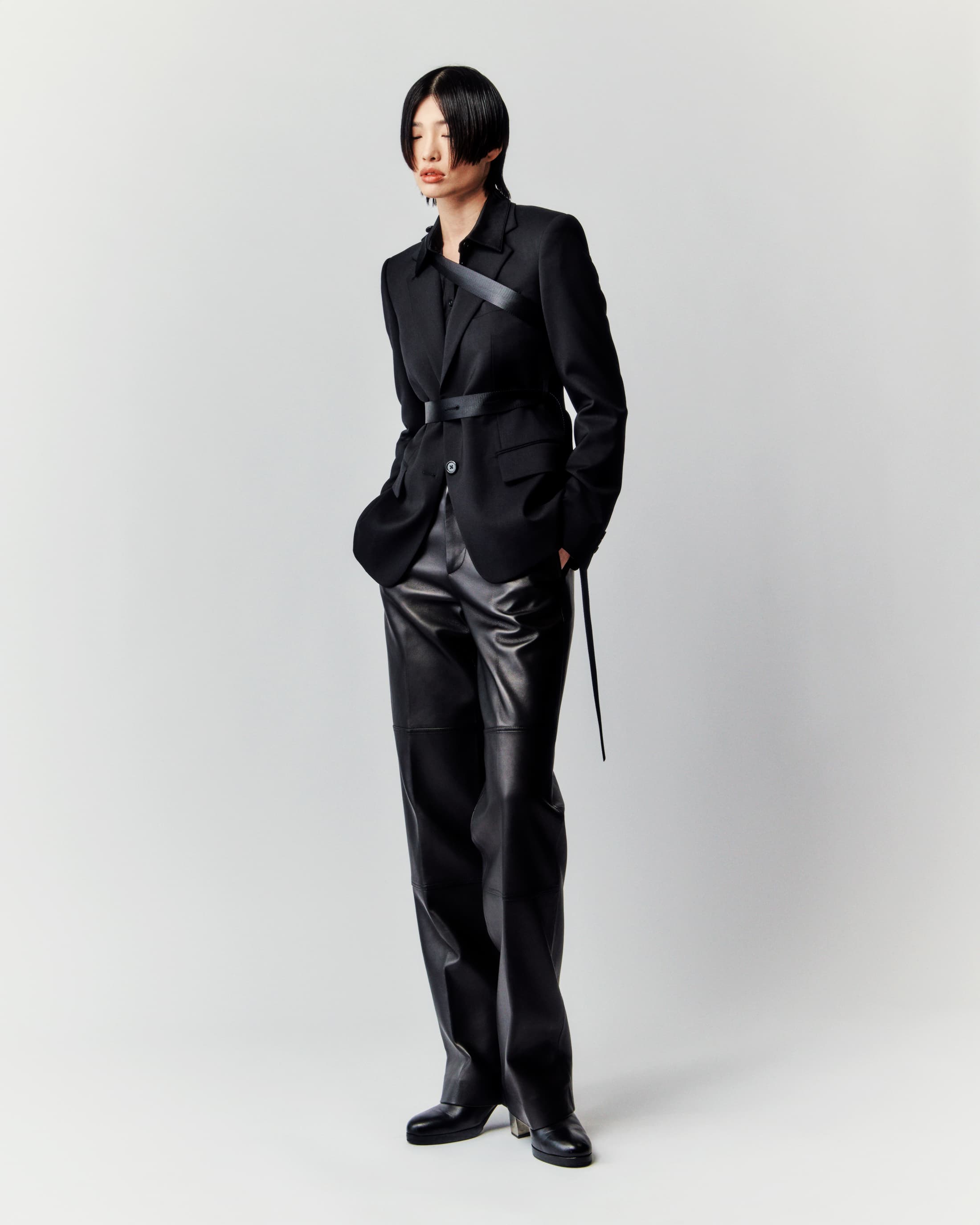A full-length studio shot presents a slender Asian woman modeling a black blazer, a belt, and shiny black pants against a plain white background.