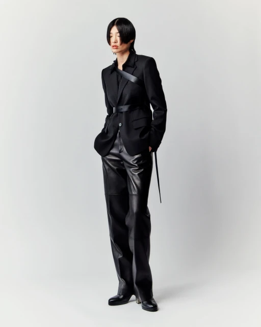 A full-length studio shot presents a slender Asian woman modeling a black blazer, a belt, and shiny black pants against a plain white background.