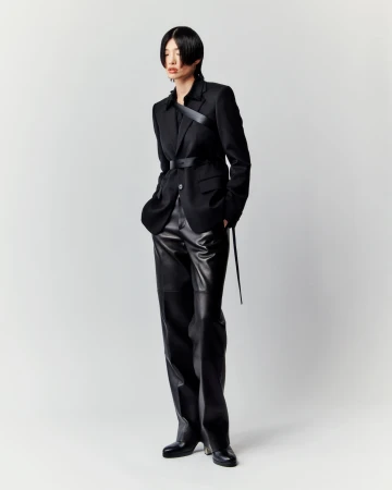A full-length studio shot presents a slender Asian woman modeling a black blazer, a belt, and shiny black pants against a plain white background.