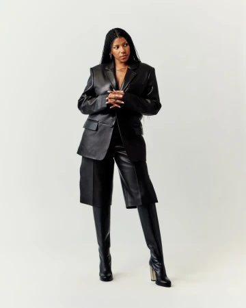 Full-length shot of an adult female with long braids wearing a black leather jacket, black leather shorts, and black thigh-high leather boots.