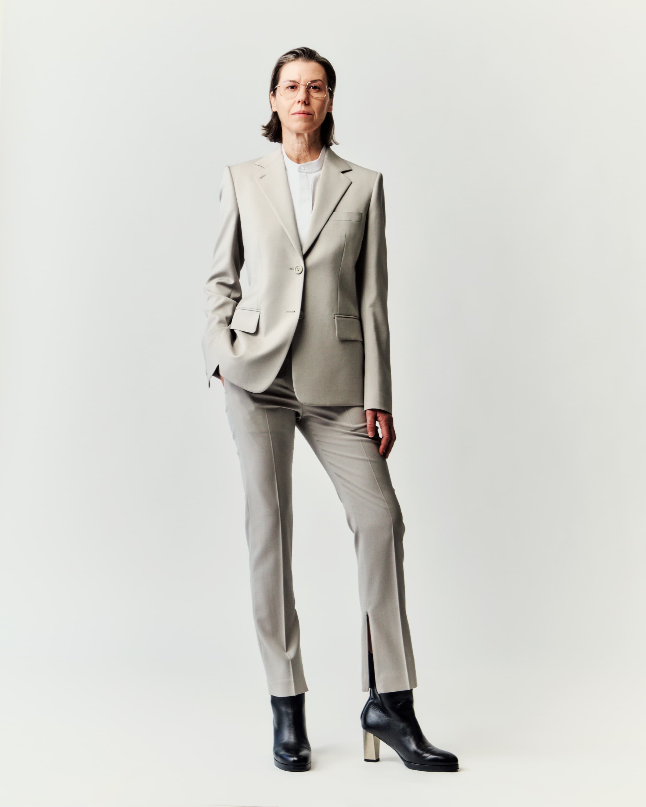 Full-length studio portrait of an adult woman with short brown hair wearing a tailored light-grey suit and black ankle boots.