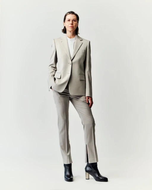 Full-length studio portrait of an adult woman with short brown hair wearing a tailored light-grey suit and black ankle boots.