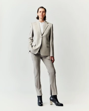 Full-length studio portrait of an adult woman with short brown hair wearing a tailored light-grey suit and black ankle boots.