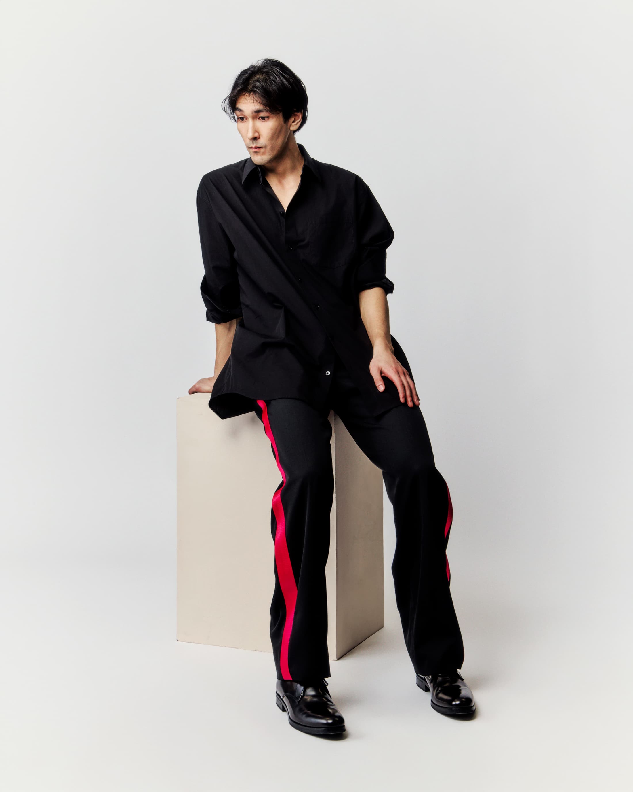 An Asian male model in a black shirt and black pants with red side stripes poses while sitting on a cream-colored cube against a white background.