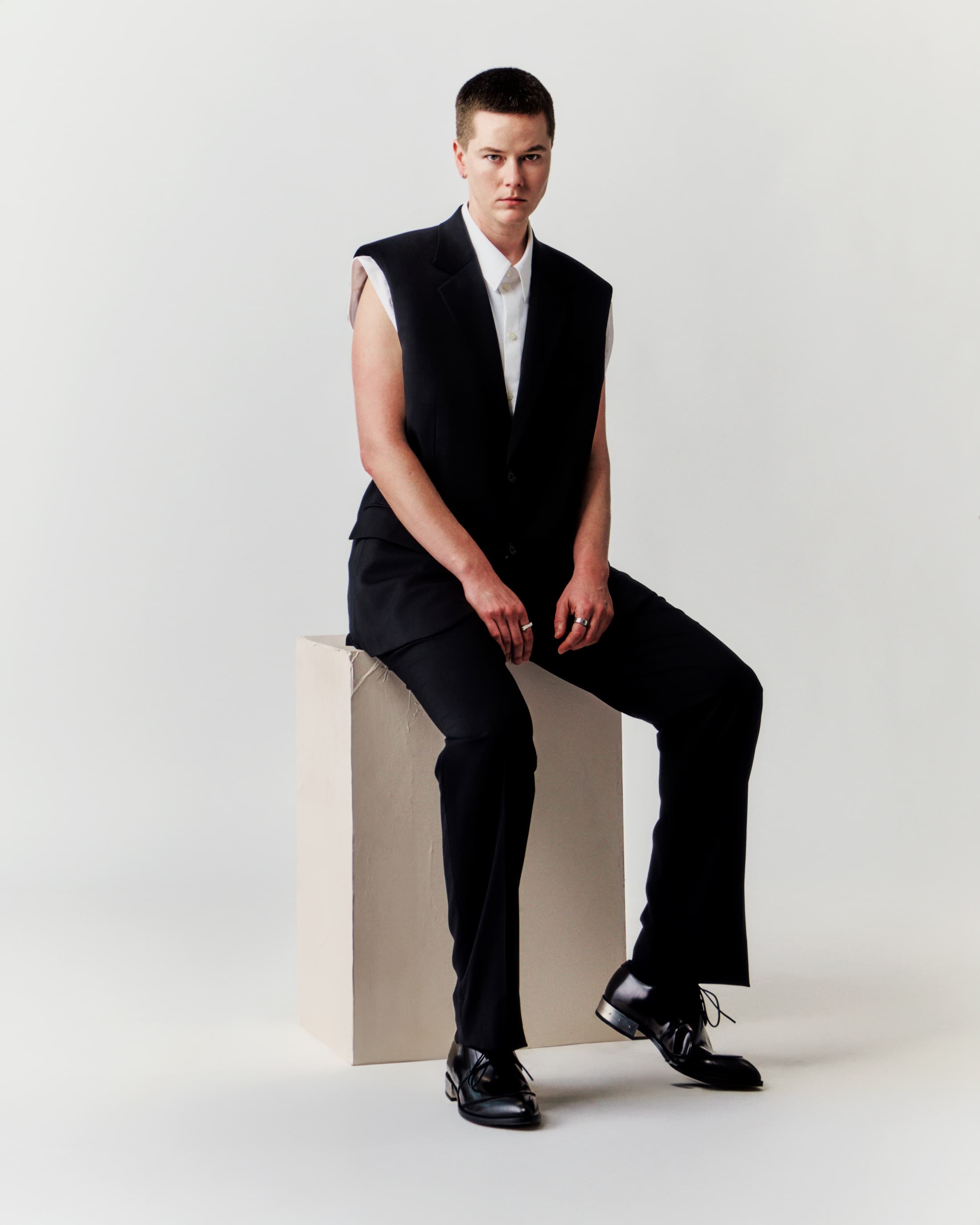 Portrait of a person with short hair in a black vest and pants sitting on a beige cube in a white photo studio setting.