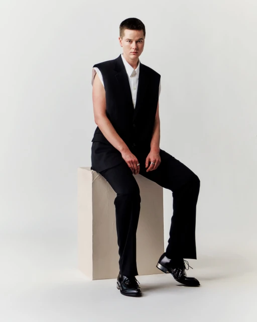 Portrait of a person with short hair in a black vest and pants sitting on a beige cube in a white photo studio setting.