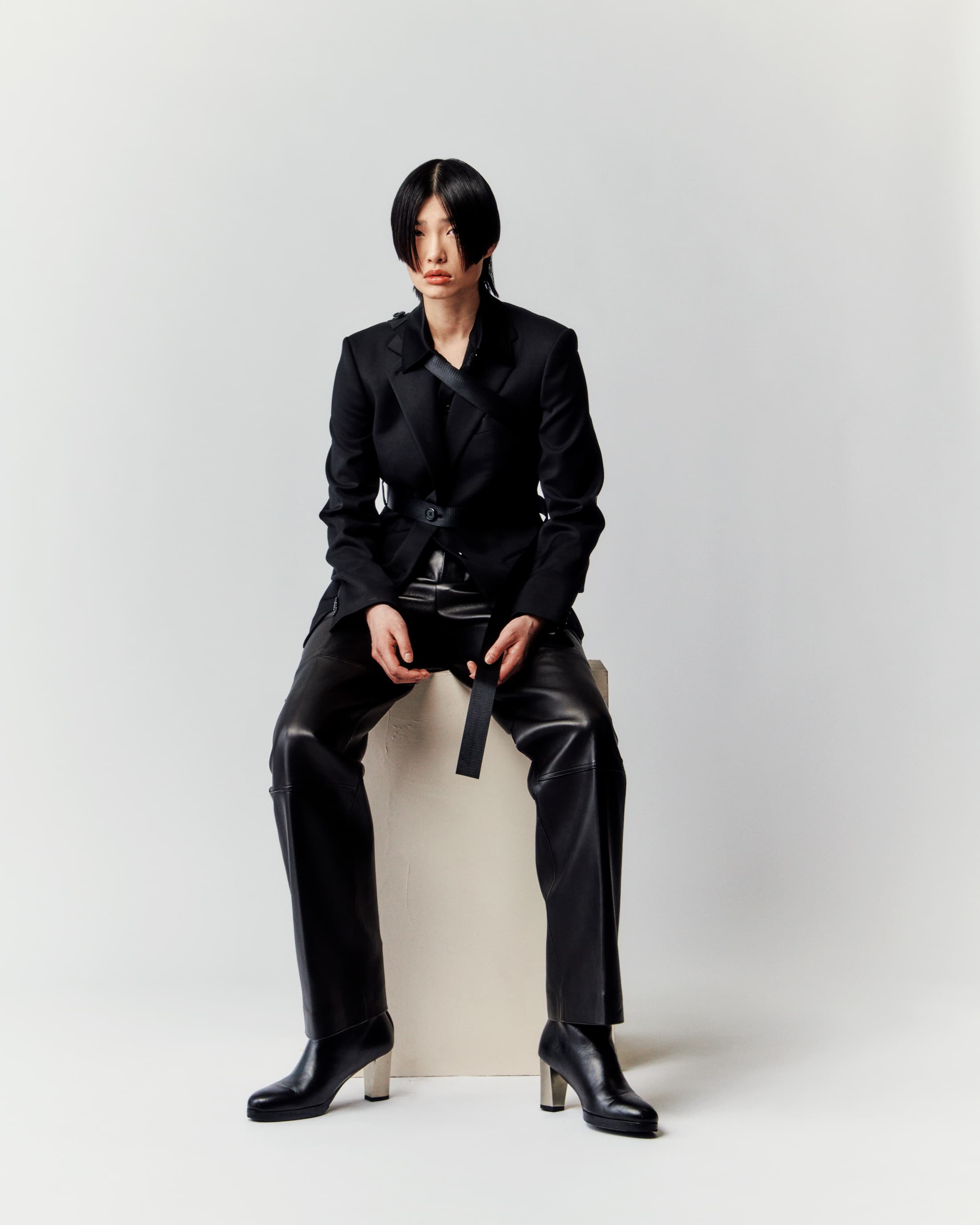 Asian woman in a black blazer and leather pants sitting on a white block with a neutral expression in a photo studio.