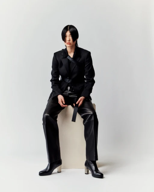 Asian woman in a black blazer and leather pants sitting on a white block with a neutral expression in a photo studio.