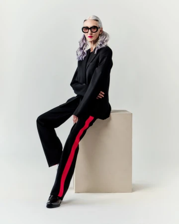 An older woman with silver hair wearing a black blazer and trousers with red side stripes sits on a cube against a white background.