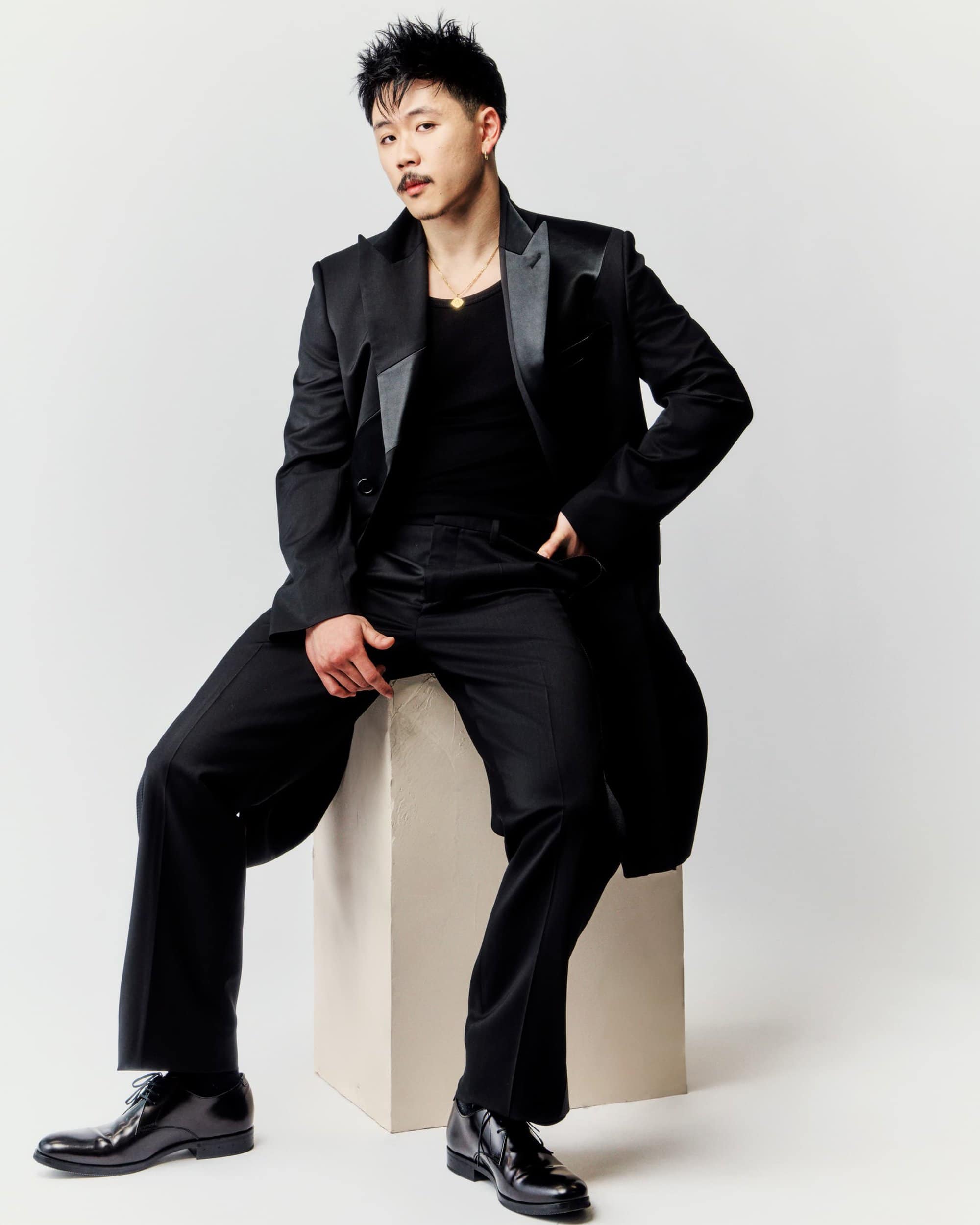 Portrait of a young adult man in black suit sitting on concrete block in a photo studio setting with a neutral background.