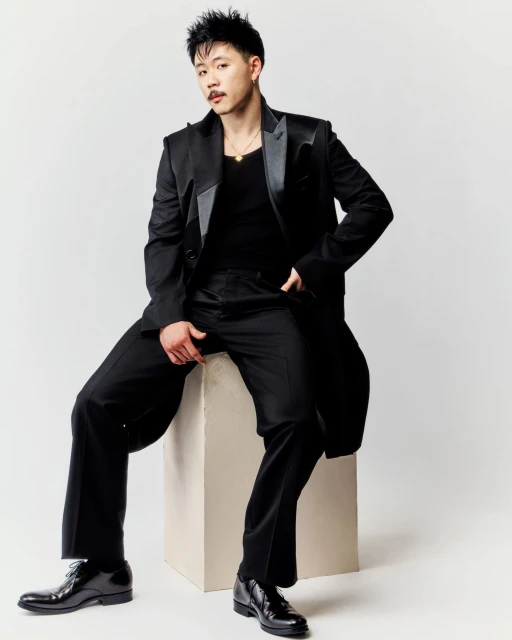 Portrait of a young adult man in black suit sitting on concrete block in a photo studio setting with a neutral background.