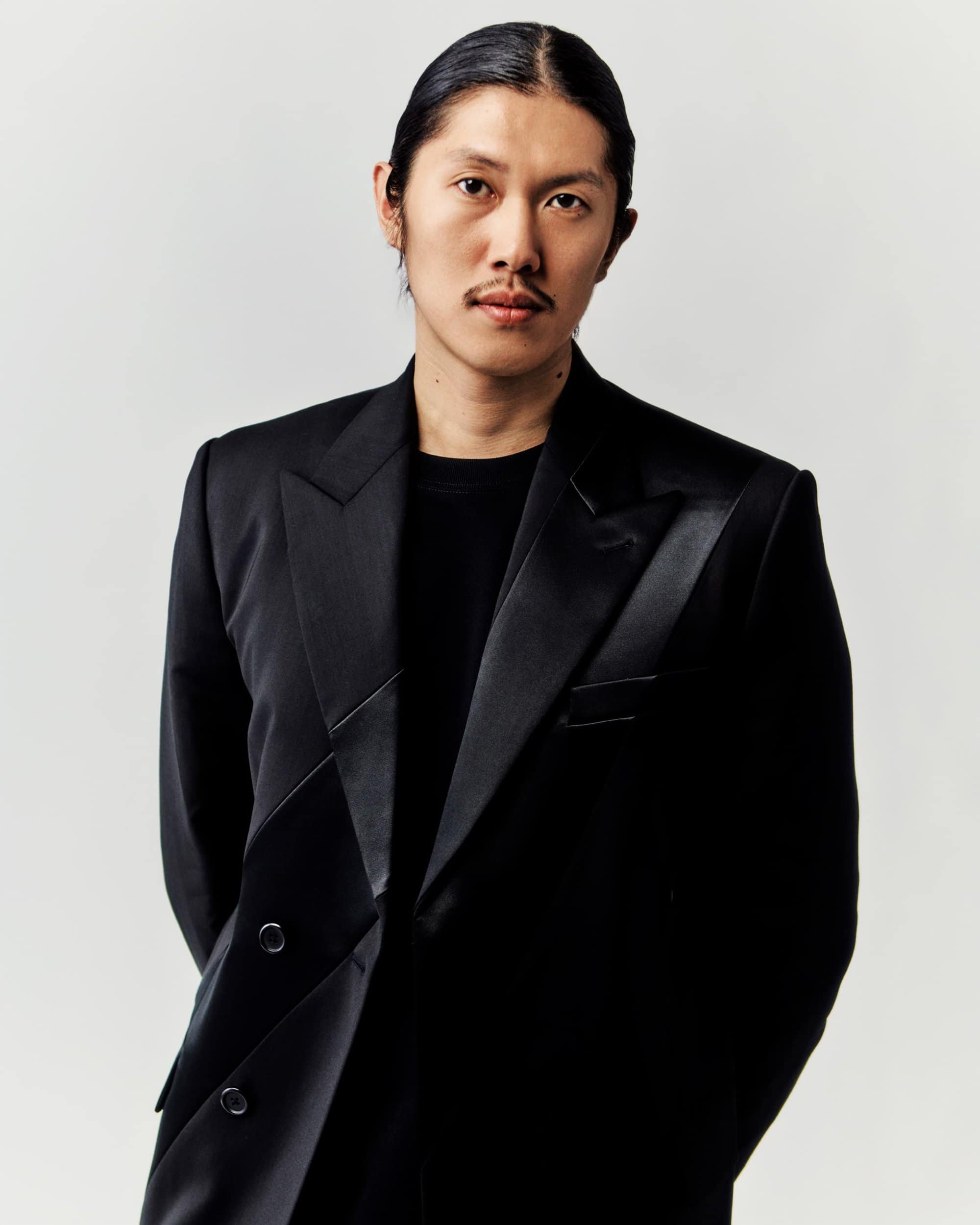Asian man in a formal black suit jacket and black shirt stands indoors against a bright white backdrop in a medium studio portrait.