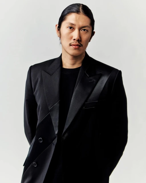 Asian man in a formal black suit jacket and black shirt stands indoors against a bright white backdrop in a medium studio portrait.