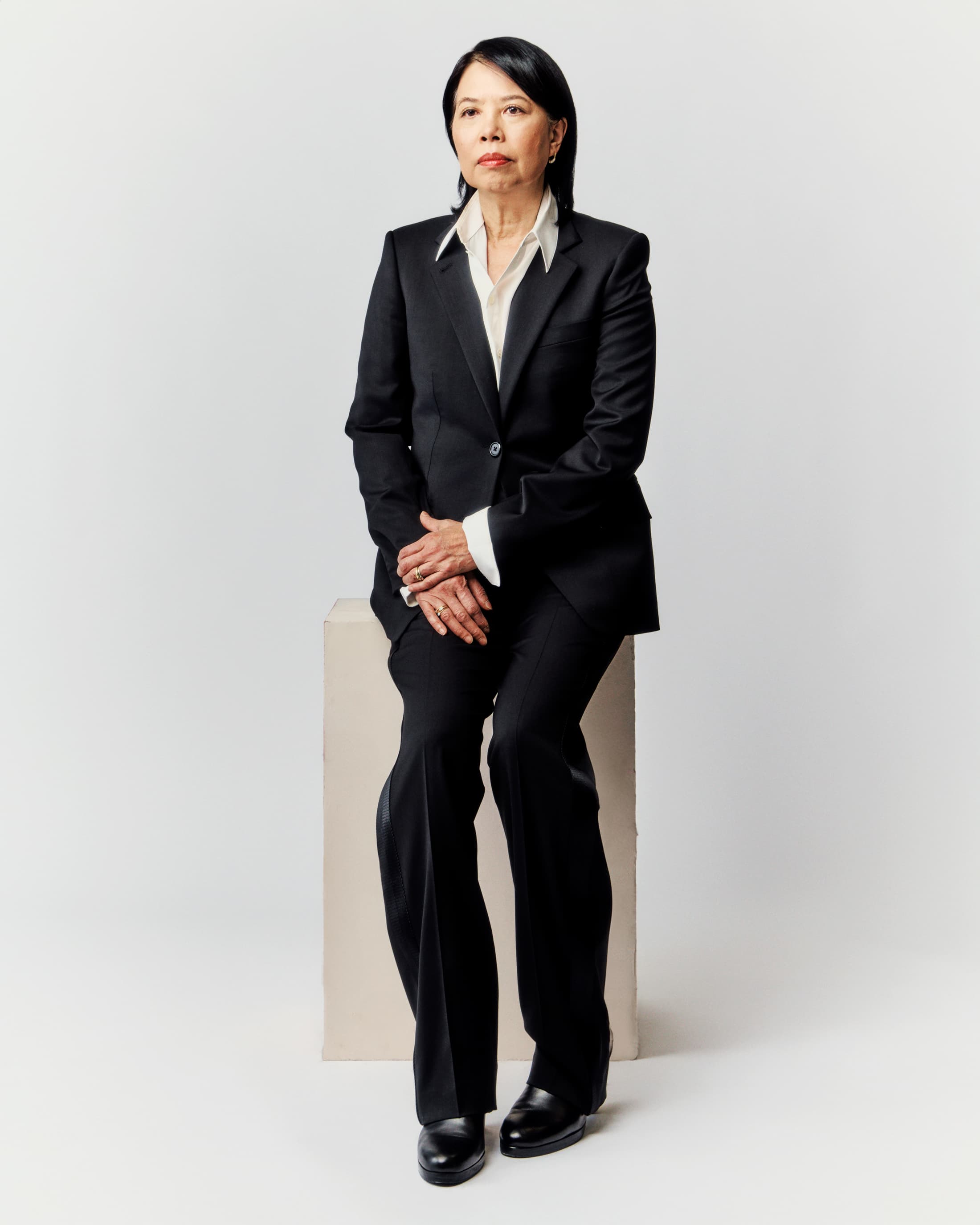 Portrait of a mature Asian woman in a black suit seated on a beige cube against a white backdrop, looking thoughtfully forward.