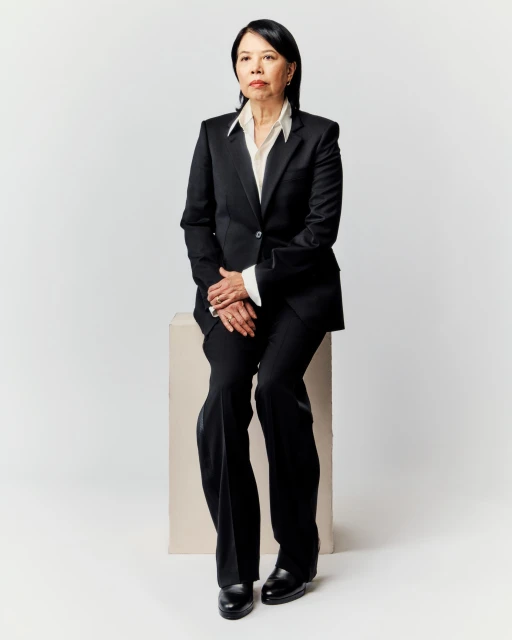 Portrait of a mature Asian woman in a black suit seated on a beige cube against a white backdrop, looking thoughtfully forward.