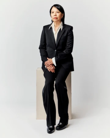 Portrait of a mature Asian woman in a black suit seated on a beige cube against a white backdrop, looking thoughtfully forward.