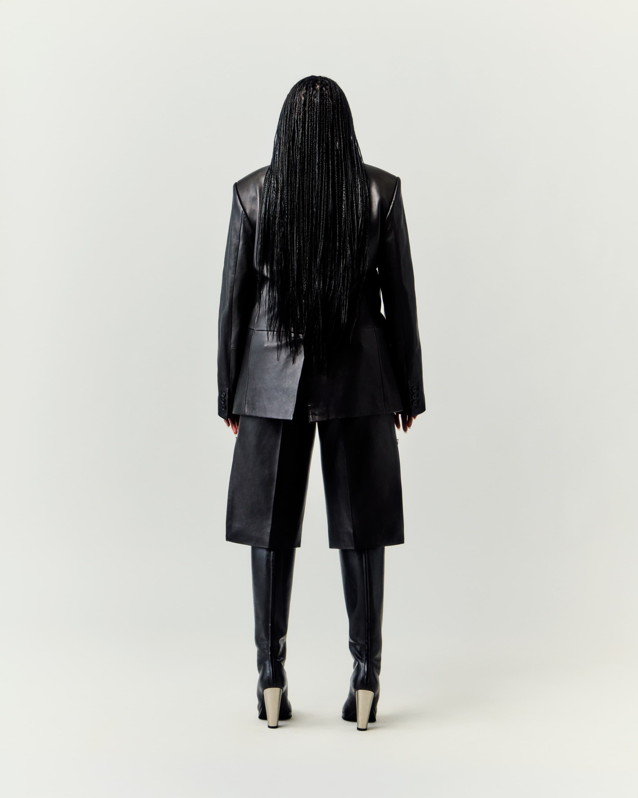 Back view of adult woman wearing black leather jacket, culottes, boots, and long braids in a bright neutral studio setting.
