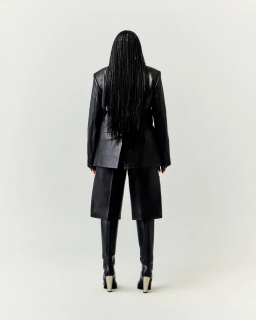 Back view of adult woman wearing black leather jacket, culottes, boots, and long braids in a bright neutral studio setting.