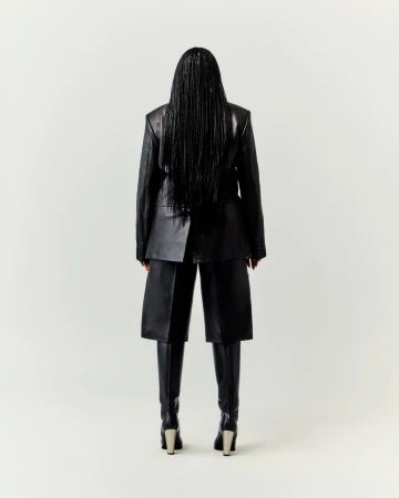 Back view of adult woman wearing black leather jacket, culottes, boots, and long braids in a bright neutral studio setting.