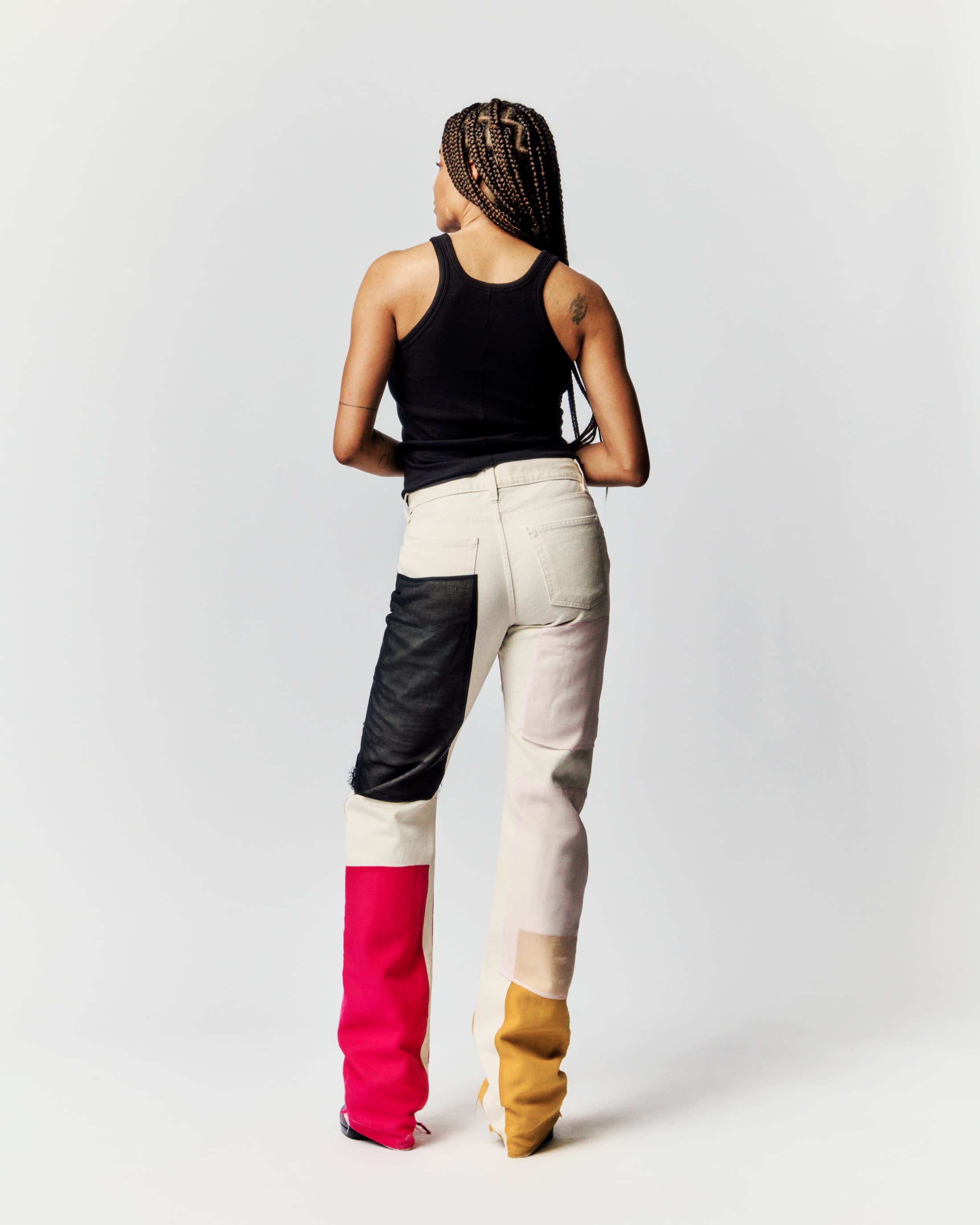 A woman stands facing away from the camera, showcasing a black tank top and color-blocked jeans with leather, pink, and tan panels, against a white background.