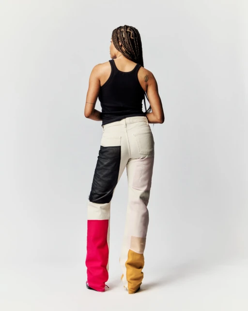 A woman stands facing away from the camera, showcasing a black tank top and color-blocked jeans with leather, pink, and tan panels, against a white background.
