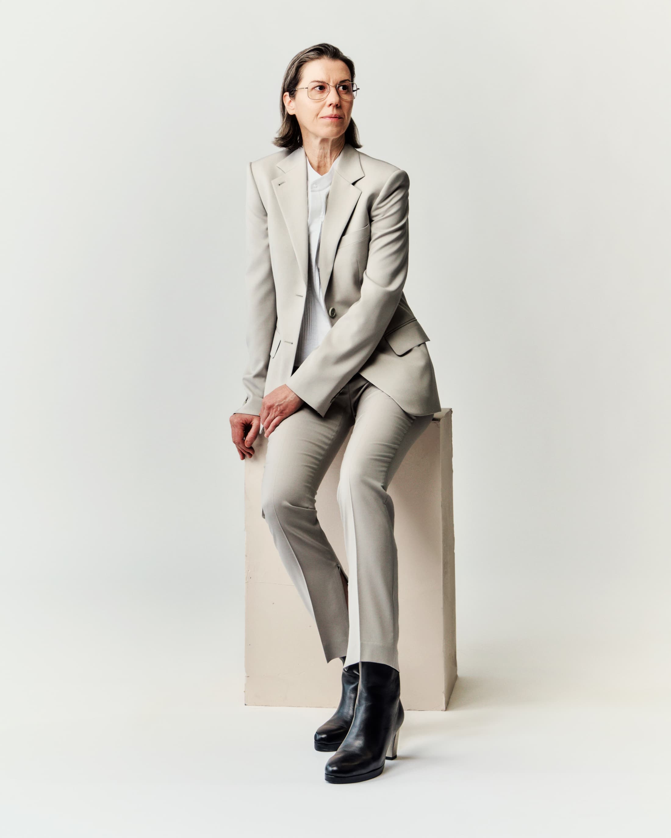 Woman in a light grey tailored pantsuit and black boots seated on a block against a plain white background.