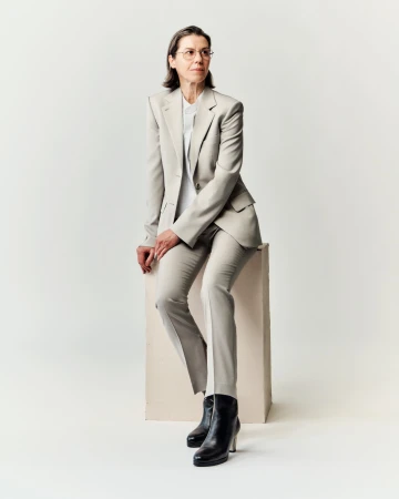 Woman in a light grey tailored pantsuit and black boots seated on a block against a plain white background.