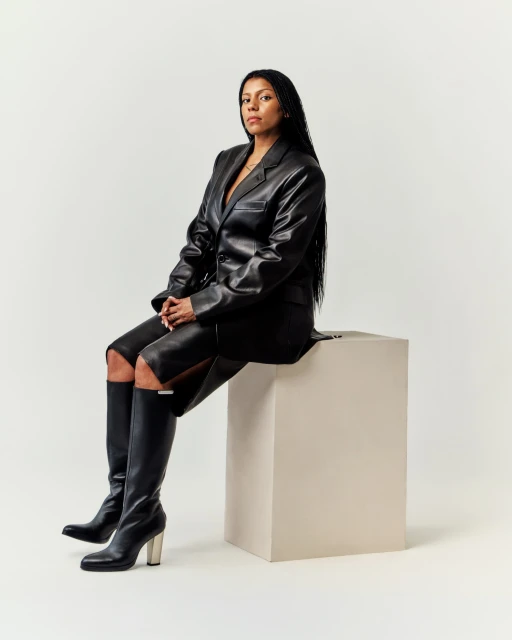 Portrait of a Black woman in a black leather jacket and skirt, seated on a white cube in a white studio with knee-high boots.