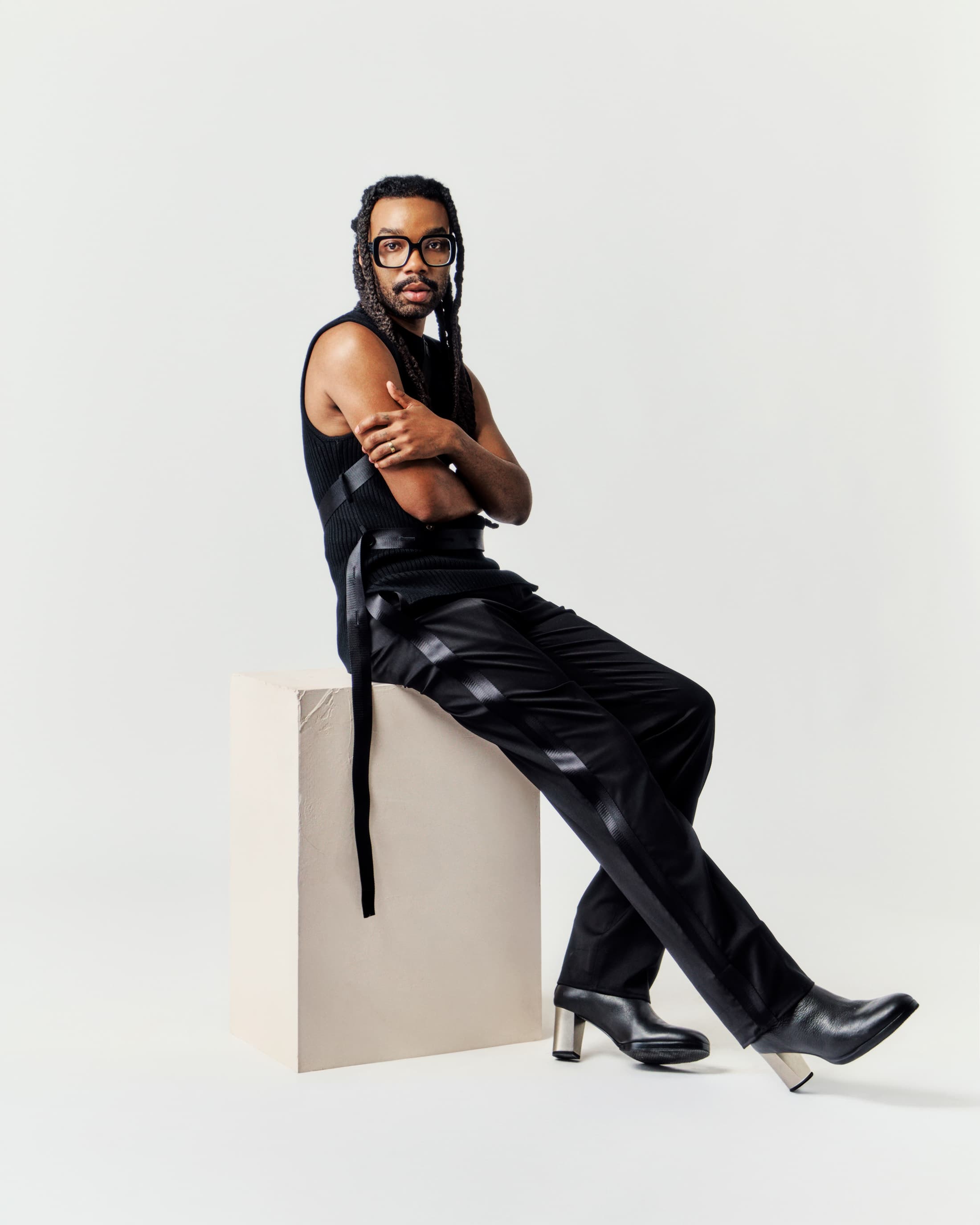 An androgynous person with long braids, wearing black vest and pants with silver boots, sits on a white cube in a bright photo studio.