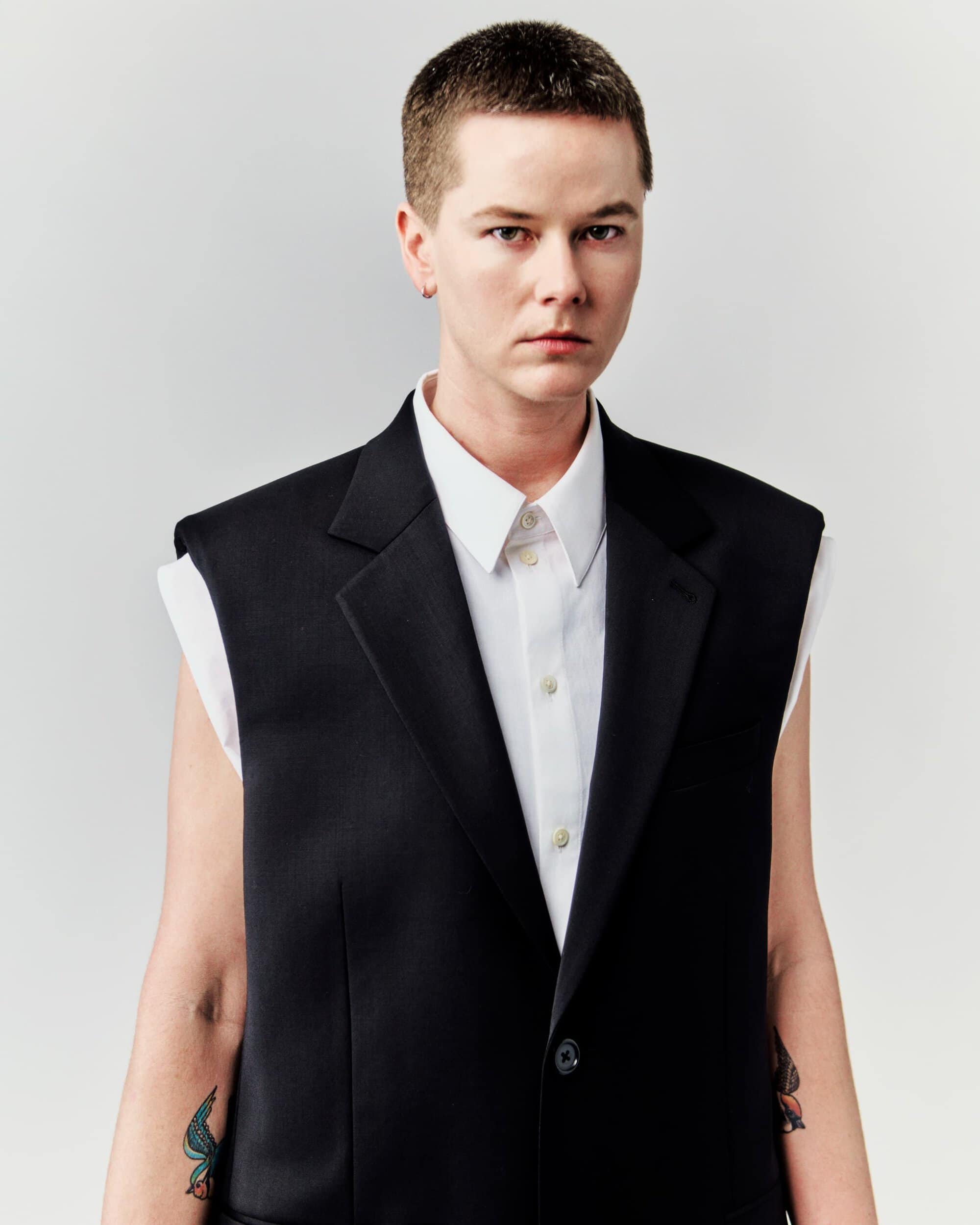 Portrait of a person with short hair wearing a black vest and white shirt against a neutral background in an indoor setting.