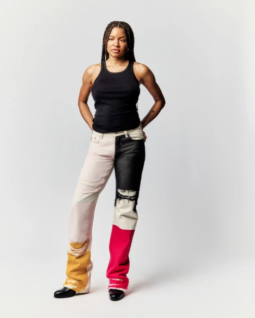 Full-length studio shot of an adult woman in a black tank top and multicolored denim pants against a neutral background, hands on hips.