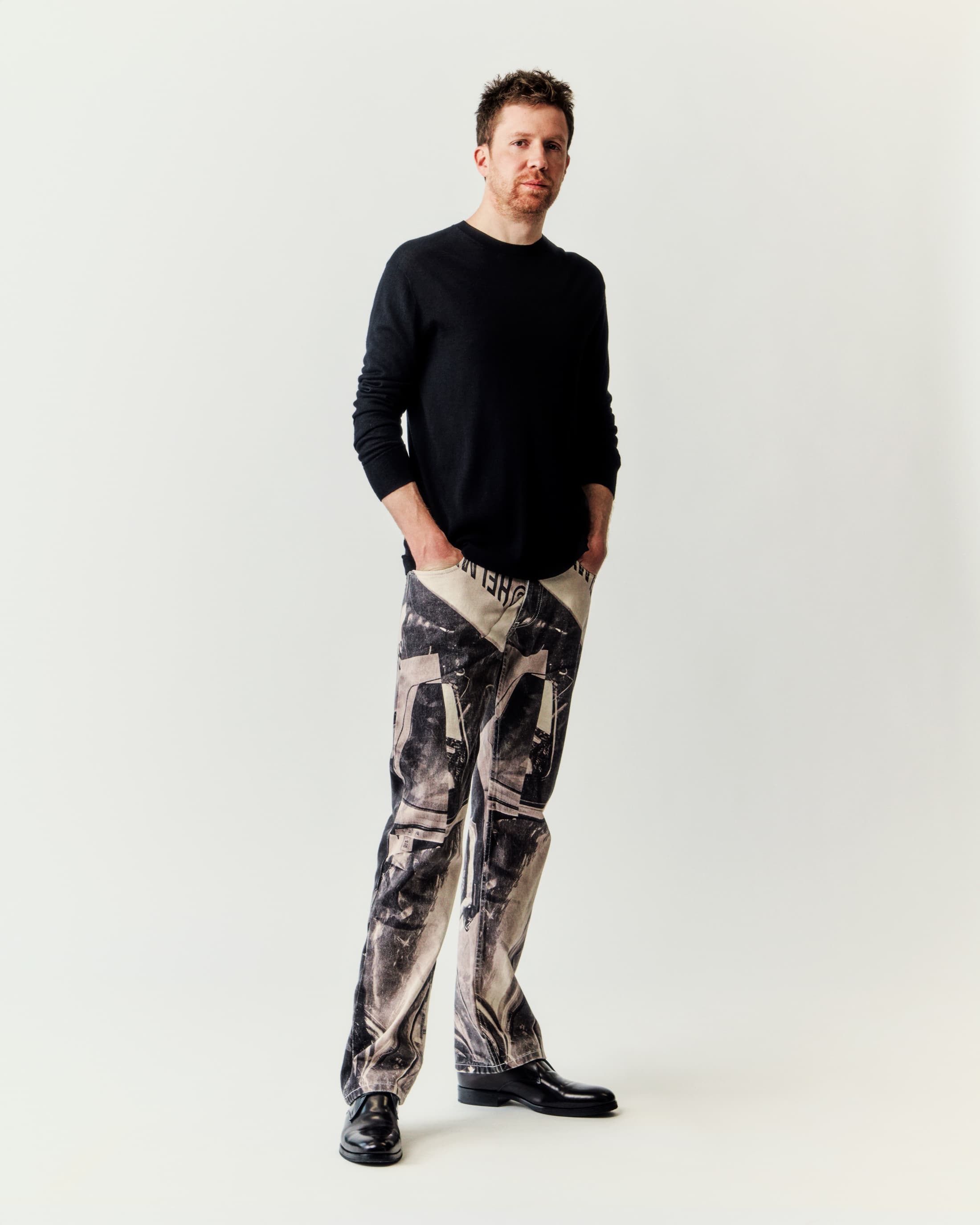 Portrait of a red-haired man wearing printed jeans, a black sweater, and black shoes, standing in front of a white background.