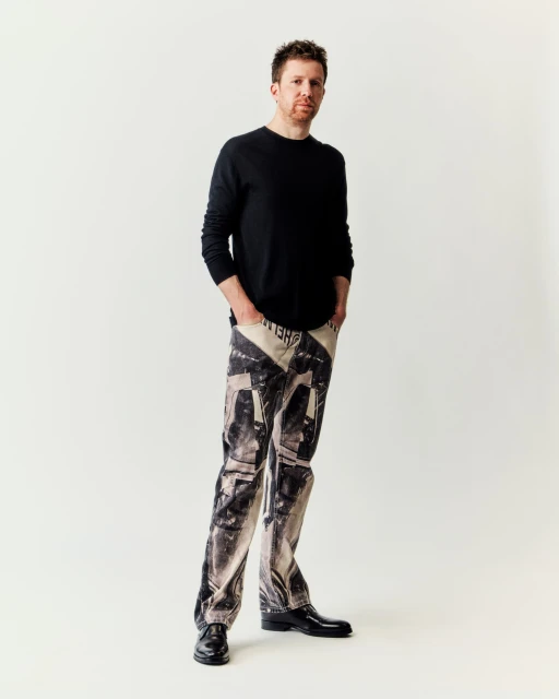 Portrait of a red-haired man wearing printed jeans, a black sweater, and black shoes, standing in front of a white background.