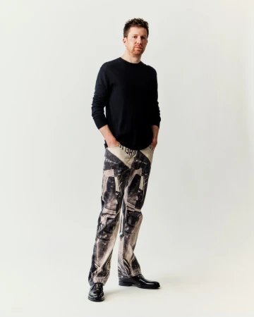Portrait of a red-haired man wearing printed jeans, a black sweater, and black shoes, standing in front of a white background.