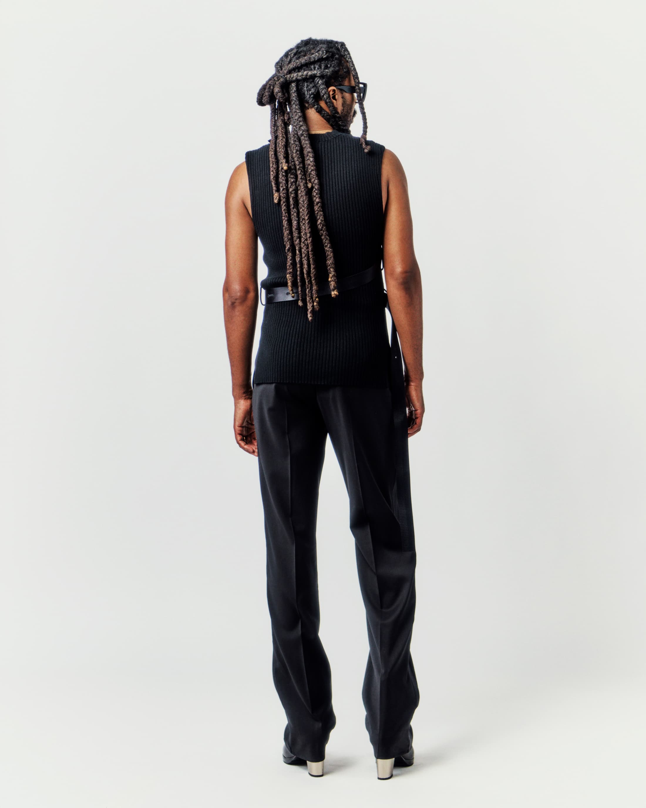 Back view of an adult with dreadlocks wearing a black vest and black dress pants against a light neutral backdrop in a studio setting.