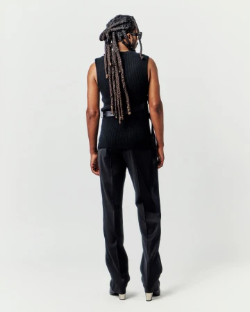 Back view of an adult with dreadlocks wearing a black vest and black dress pants against a light neutral backdrop in a studio setting.
