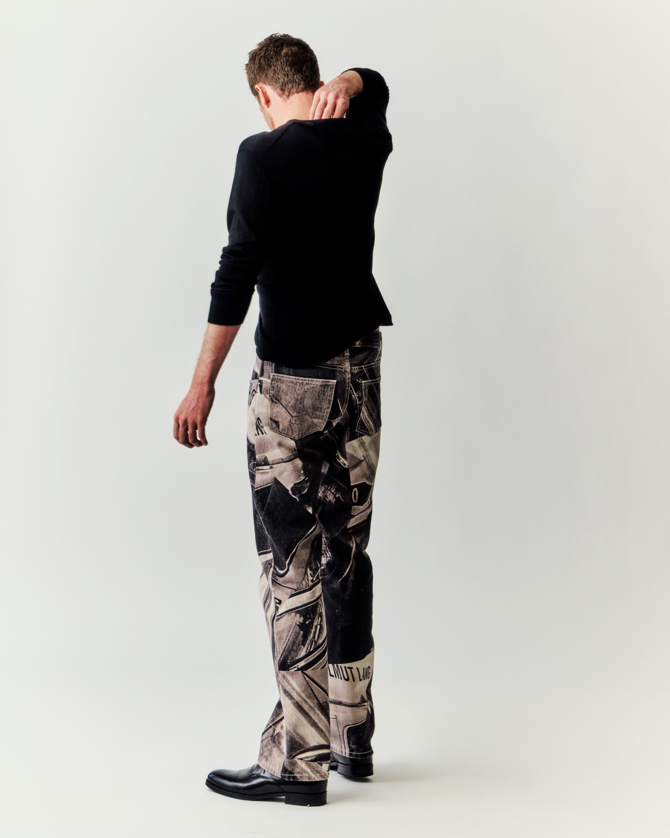 Adult male in black long-sleeved shirt and printed denim stands with back to the camera in a brightly lit photo studio.