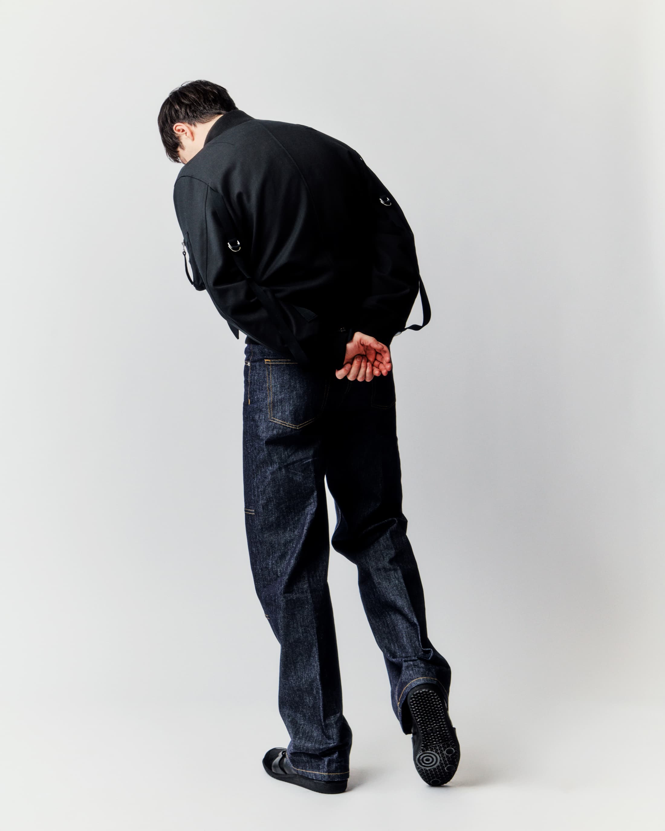 Back view of an adult man wearing a black jacket, dark jeans, and casual shoes, posing in a brightly lit photo studio against a white backdrop.