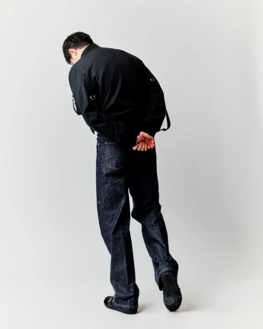 Back view of an adult man wearing a black jacket, dark jeans, and casual shoes, posing in a brightly lit photo studio against a white backdrop.