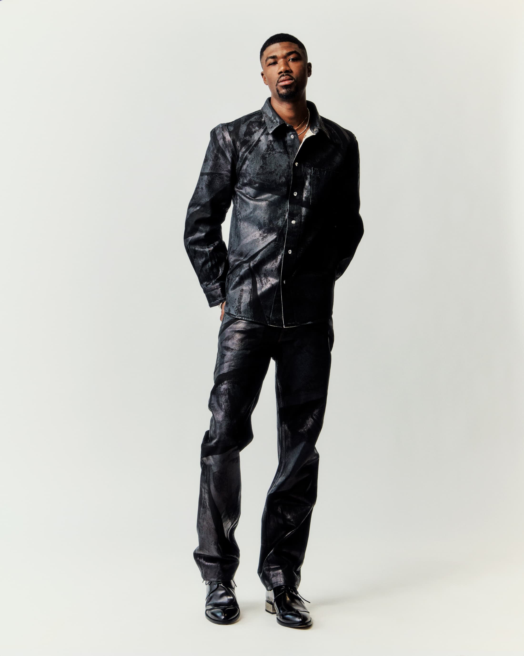 A full studio shot of a Black male model posing, wearing a black snakeskin-patterned shirt and pants with matching black shoes, against a white backdrop.
