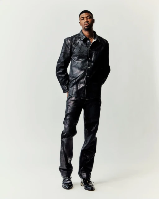 A full studio shot of a Black male model posing, wearing a black snakeskin-patterned shirt and pants with matching black shoes, against a white backdrop.