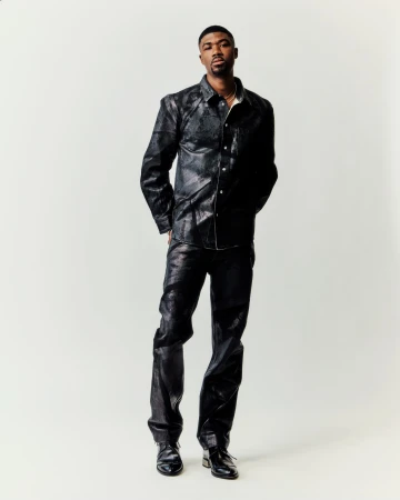 A full studio shot of a Black male model posing, wearing a black snakeskin-patterned shirt and pants with matching black shoes, against a white backdrop.