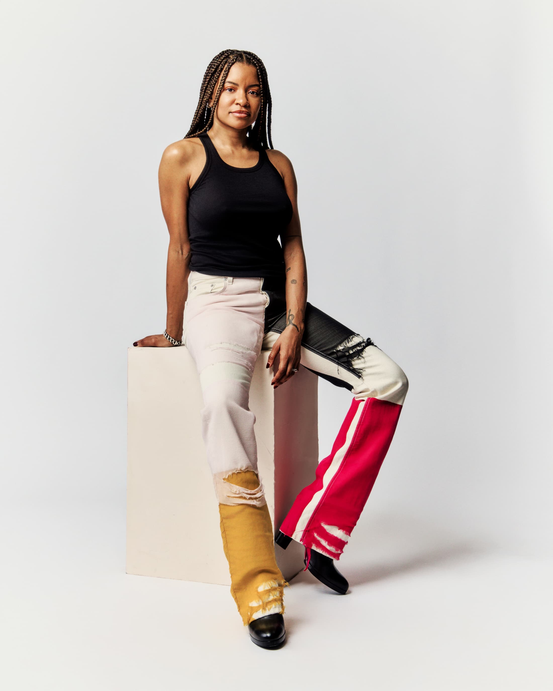 A woman with braids poses in a studio. She is wearing a black tank top and patchwork jeans while sitting on a white block.